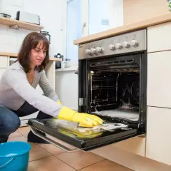 Frau mit gelben Handschuhen reinigt den Backofen. Schaum und ein blauer Eimer stehen bereit.