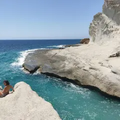 Una chica sentada en una roca en una cala chica, con el mar azul turquesa y acantilados blancos al fondo.