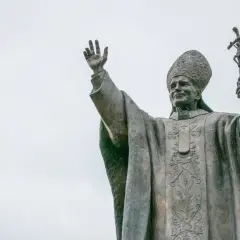 Pomnik Jana Pawła II z podniesioną ręką i krzyżem. Wierni odmawiają modlitwę do Jana Pawła II o uzdrowienie.
