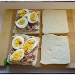 Deux sandwichs oeuf dur sont en préparation sur une planche en bois. Le thon émietté et la mayonnaise sont déjà étalés sur le pain.
