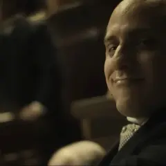 Un uomo con un sorriso furbo sembra rompere la quarta parete, guardando direttamente lo spettatore.