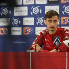 Piłkarz w czerwonej bluzie przed mikrofonami, gotowy do rozmowy o nadchodzących meczach Widzew Łódź – Lech Poznań.