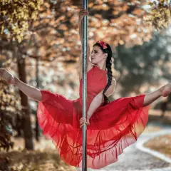 Kobieta w czerwonej sukience wykonuje figurę na słupie, jak podczas kursu pole dance. Jej nogi są rozłożone w szpagacie, a włosy splecione w warkocz.