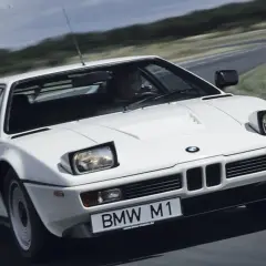 Białe BMW M1 na zakręcie drogi, z widocznym kierowcą i tablicą rejestracyjną.