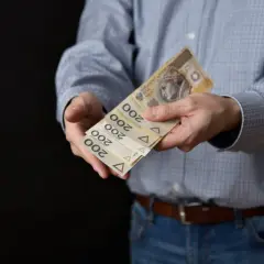 Mężczyzna w koszuli i jeansach podaje kilka banknotów po 200 zł. Może to być symbol zapłaty, darowizny lub cesji.
