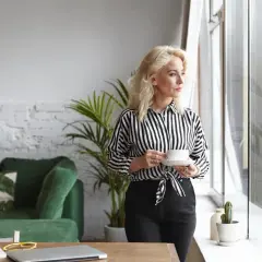 Kobieta z kawą przy oknie, zastanawia się, jak znaleźć właściciela mieszkania. W tle zielona kanapa i rośliny.