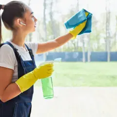 Dziewczyna w żółtych rękawiczkach i granatowym ogrodniczkach pokazuje, jak myć okna bez smug, używając niebieskiej ściereczki i butelki ze sprayem.