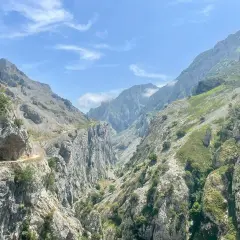 Un senderista recorre la **ruta del Cares**, un camino tallado en la roca entre imponentes montañas verdes y grises bajo un cielo azul con nubes.