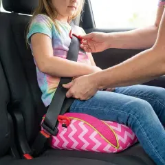 Ein Kind sitzt auf einem pinken Sitzkissen mit Zickzackmuster. Ein Erwachsener befestigt den Sicherheitsgurt, um den Kindersitz zu sichern.