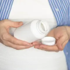Kobieta w ciąży trzyma białą buteleczkę i jej nakrętkę. Rozważa, czy wziąć paracetamol w ciąży.