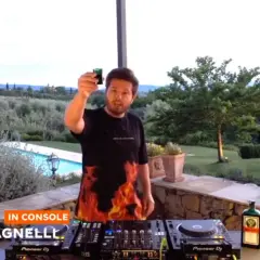 Luca Agnelli, uno dei dj techno famosi, brinda con un bicchierino davanti alla console, con una piscina e un paesaggio collinare sullo sfondo.