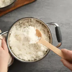 Hände halten einen Topf mit frisch gekochtem Reis. Eine hölzerne Kelle hebt eine Portion Reis heraus, perfekt auf welcher Stufe Reis kochen.