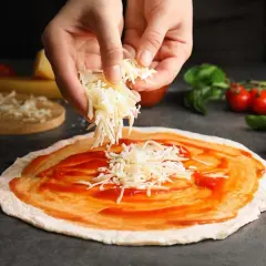 Hände streuen geriebenen Gouda auf eine mit Tomatensauce bestrichene Pizza. Tomaten und Basilikum liegen daneben.