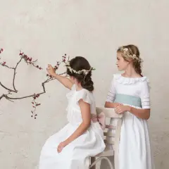 Dos niñas con peinados de comunión con diadema floral, una sentada tocando ramas, la otra de pie.
