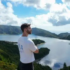 Hombre contemplando la **Lagoa do Fogo**, un lago azul rodeado de montañas verdes y un cielo nublado.