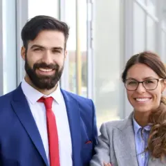 Smiling professionals, a man in a blue suit and red tie, and a woman in a grey blazer, stand outside. They're ready to pursue online MPA programs no GRE.
