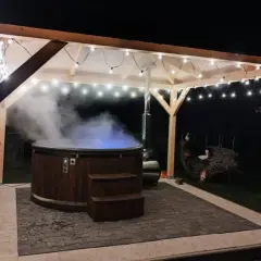 Drewniana beczka z gorącą wodą, z której unosi się para, pod zadaszeniem ozdobionym lampkami. Idealne miejsce na relaks w stylu sauna fińska.