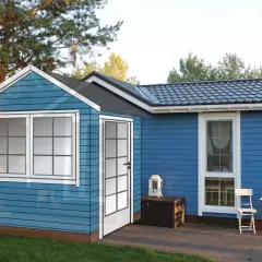 En blå stuga med vita fönster och dörr, redo att bygga farstukvist. En liten uteplats med bord och stolar.