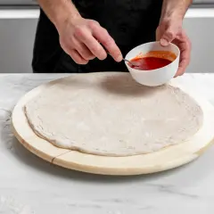 Ręce kucharza przygotowują szybką pizzę, nabierając sos łyżeczką z miseczki na surowe ciasto.
