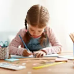 Dziewczynka z warkoczami pisze kredką w zeszycie. Na biurku kolorowe kredki, nożyczki i stos książek. Ćwiczenia grafomotoryczne cele to rozwój precyzji ruchów.