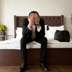 Zmartwiony mężczyzna w garniturze siedzi na łóżku, zakrywając twarz dłońmi. Czy właściciel może wyrzucić z mieszkania? Obok torba podróżna.