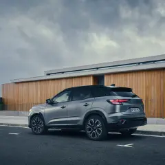 Vue arrière d'un SUV gris Renault Austral, stationné sur une route devant un bâtiment moderne en bois. La motorisation est performante.