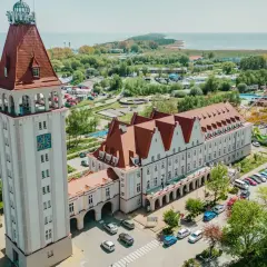 Wieża zegarowa i długi budynek z czerwonym dachem, przypominający dom rybaka, stoją obok siebie. W tle widać morze i park rozrywki.