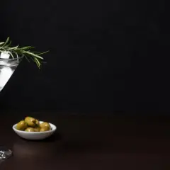 Elegancki drink z oliwką i gałązką rozmarynu, obok miseczka z zielonymi oliwkami.