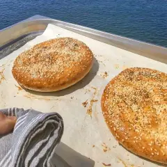 Två gyllene ramazan pidesi, toppade med sesamfrön, vilar på ett bakplåtspapper vid havet.