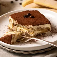 Ein Stück Tiramisu, zubereitet nach einem einfachen tiramisu rezept, liegt auf einem Teller. Kakaopulver bestäubt die cremige Schicht und die Löffelbiskuits.