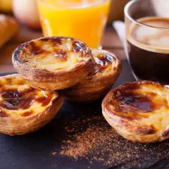 Pyszne babeczki portugalskie z kremowym nadzieniem i karmelizowaną skórką, obok kawy i soku pomarańczowego.