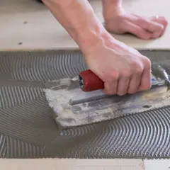 A hand uses a notched trowel to spread thinset for cement board, creating a textured surface for tile adhesion.