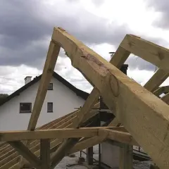 Montaż więźby dachowej w toku. Drewniane belki tworzą konstrukcję dachu nad białym domem z kominem.