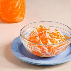 A bowl of vibrant japanese pickled carrots and daikon radish sits on a blue plate, with a jar of the same pickled vegetables in the background.