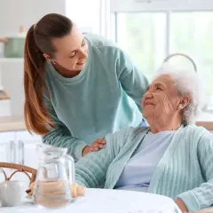 Młoda kobieta wspiera starszą panią przy stole. Rozmowa o tym, ile wynosi zasiłek opiekuńczy, jest ważna dla zapewnienia godnego życia seniorom.