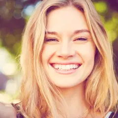 Strahlendes Lächeln einer jungen Frau mit blonden Haaren, die eine natürliche Frisur trägt.