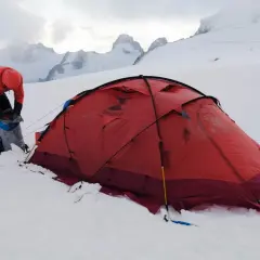 Osoba w pomarańczowej kurtce przygotowuje sprzęt przy czerwonym namiocie. W tle majestatyczne, ośnieżone szczyty, zapowiadające trudne wejście na Mount Everest.