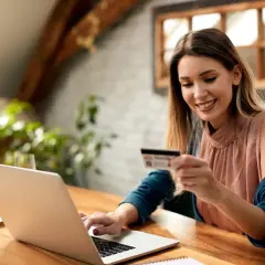 Uśmiechnięta kobieta z kartą płatniczą przy laptopie, planuje zakupy. Sprawdza cenę netto przed finalizacją transakcji.
