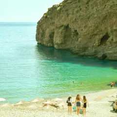 Idealna grecja pogoda lipiec: turkusowa woda, skaliste klify i plaża z parasolami. Trzy dziewczyny idą w stronę morza.