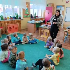Kobieta w panterkowej kurtce stoi przed grupą dzieci siedzących w kręgu na zielonej wykładzinie.