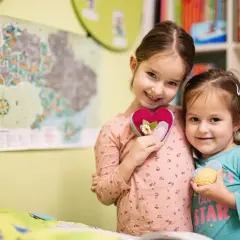 Dwie uśmiechnięte dziewczynki trzymają serduszka. Starsza w różowej bluzce, młodsza w turkusowej. W tle mapa i półka z książkami. Ciekawi ich, ile godzin w zerówce spędzają na zabawie.