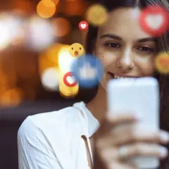 Uśmiechnięta kobieta z telefonem, otoczona emotikonami. Czy zielone serce oznacza coś więcej niż tylko polubienie?