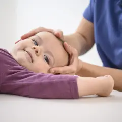 Sanfte Hände einer Osteopathin untersuchen den Kopf eines lächelnden Kleinkindes.