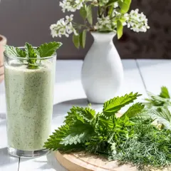 Zielony koktajl z pokrzywy, ozdobiony miętą, obok świeżych ziół na desce. Pesto z pokrzywy w szklance.