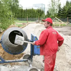 Budowlaniec w czerwonym ubraniu obsługuje betoniarkę na placu budowy, obok sterty piasku.