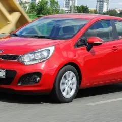 Czerwony Kia Rio 3 w ruchu, z widocznym numerem rejestracyjnym WN 7191C, na tle mostu i budynków.