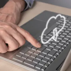 Dłoń naciska klawisz na laptopie, nad którym unosi się symbol chmury internetowej z ikoną pobierania.
