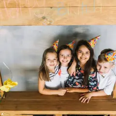 Dzieci w czapkach urodzinowych pozują przy drewnianej konstrukcji z napisem "Happy Birthday". To świetny pomysł, jak zrobić fotobudkę z kartonu na przyjęcie!