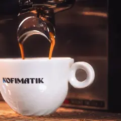 Gorąca ristretto kawa spływa do białej filiżanki z logo Kofimatik. Intensywny aromat i głęboki smak.