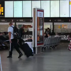 Dworzec autobusowy z tablicami odjazdów, kasami biletowymi i ludźmi czekającymi na podróż.